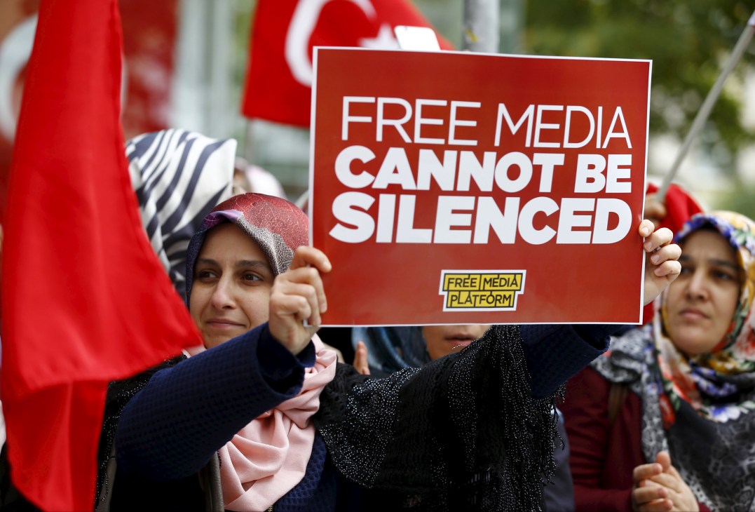Woman holding red sign reading 'FREE MEDIA CANNOT BE SILENCED' at protest with Turkish flags in background