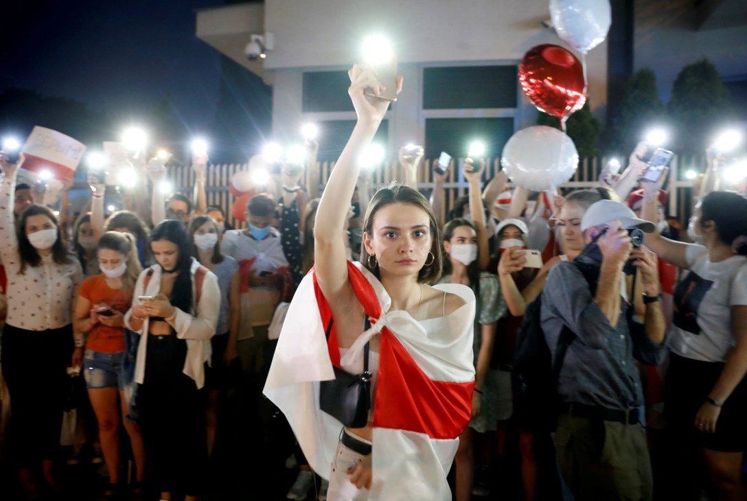 Young woman wrapped in red and white flag raises fist at nighttime protest with crowd holding phone lights and balloons