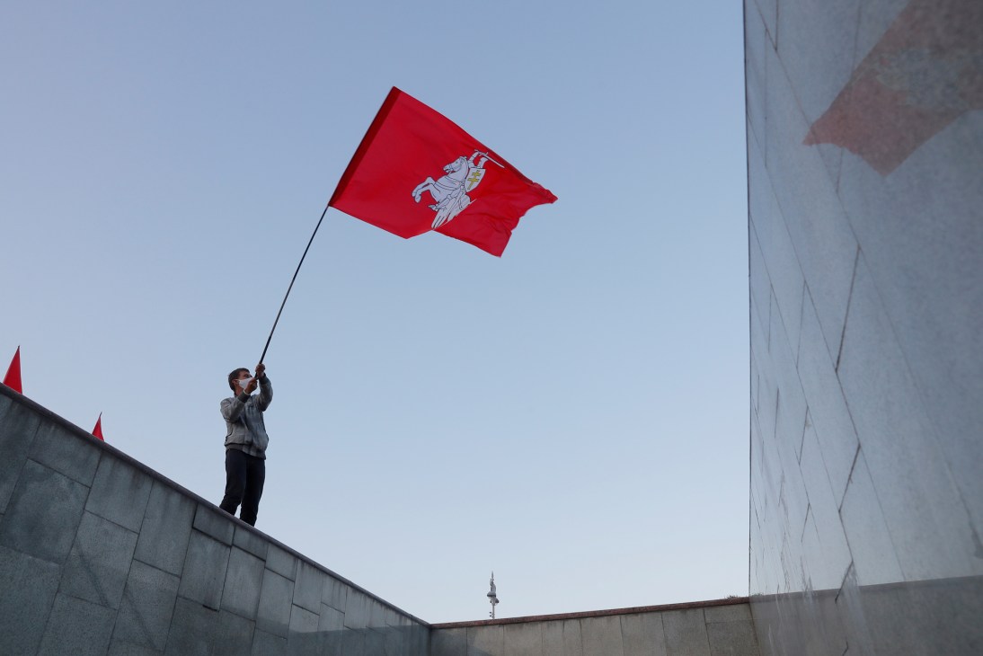 Person holding large red Belarusian flag with white knight symbol on concrete structure against blue sky