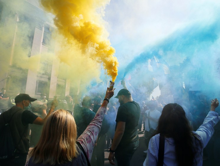 Protesters holding yellow and blue smoke flares during a demonstration on a city street.