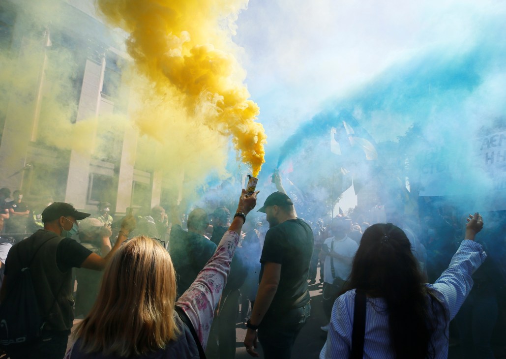 Protesters holding yellow and blue smoke flares during a demonstration on a city street.