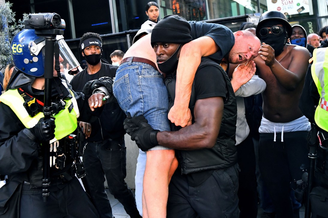 Black man in dark clothing carrying injured white man over his shoulder during street protest