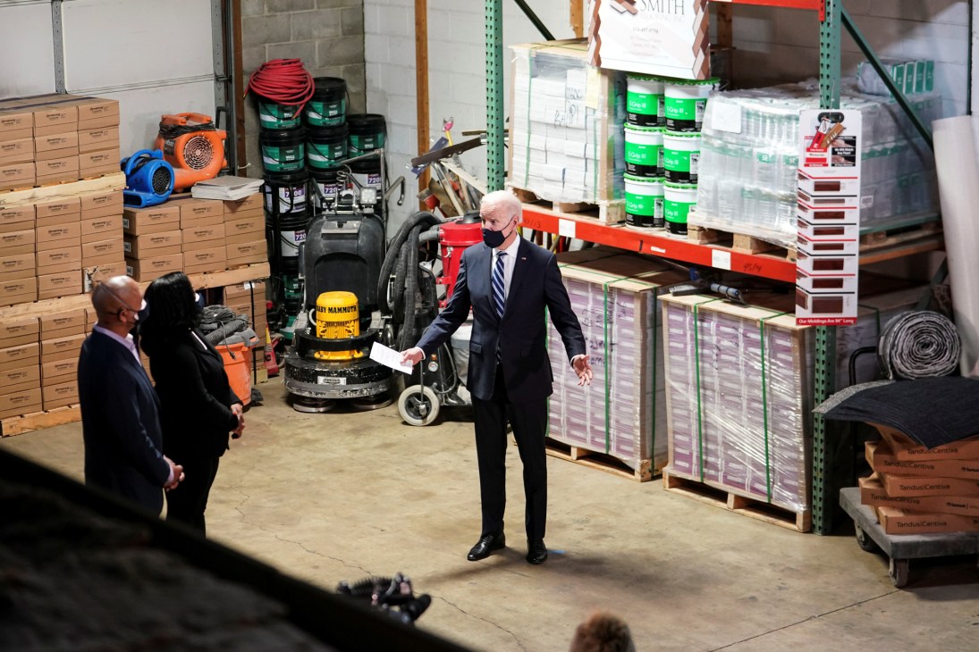 Biden speaking at a podium in a warehouse setting with supplies and shelving visible in the background