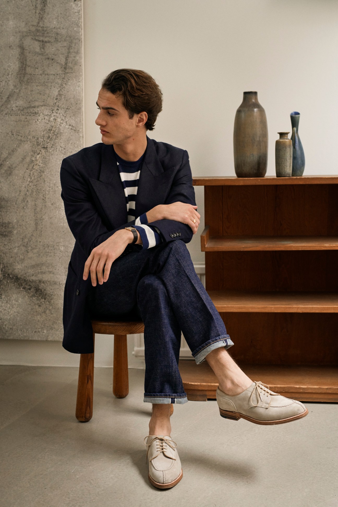 Man sitting on wooden chair wearing navy blazer, striped shirt, and cream lace-up oxfords, with rolled denim jeans, against minimalist interior backdrop with ceramic vessels on shelf.
