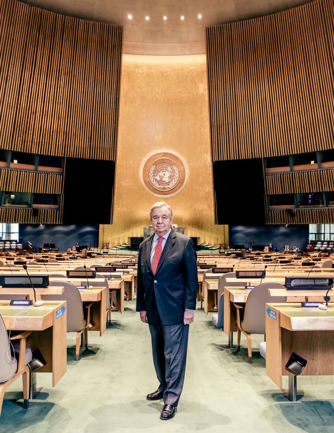 António Guterres standing in the empty UN General Assembly hall with wooden desks and the UN emblem visible