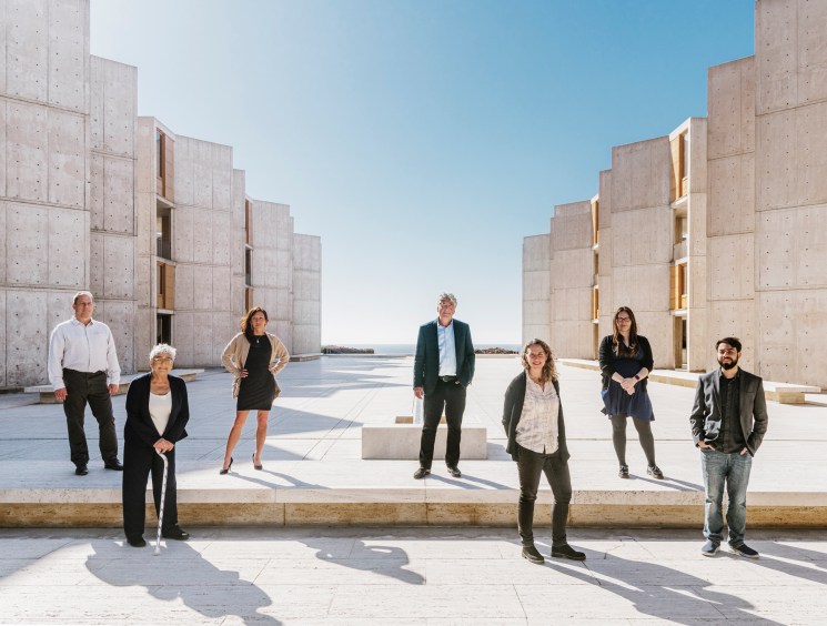 Six researchers standing spaced apart in the concrete courtyard of the Salk Institute's modernist building