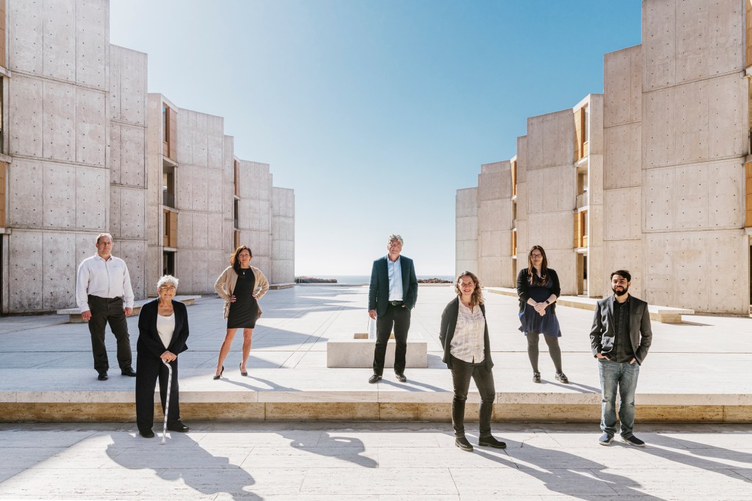 Six researchers standing spaced apart in the concrete courtyard of the Salk Institute's modernist building