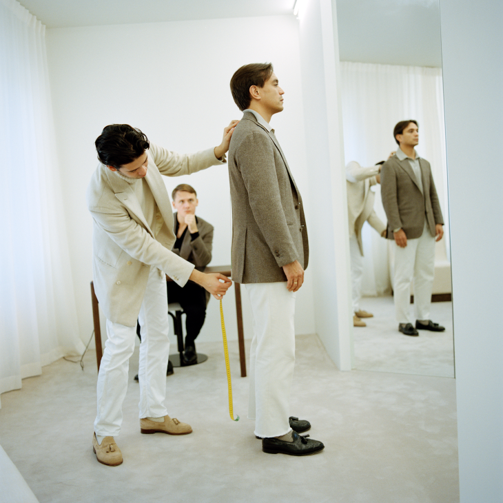 A tailor measures a client's shoulders in a minimalist fitting room with neutral tones and a mirror.