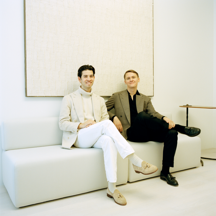 Two men in casual attire sitting on a white sofa against a plain wall.