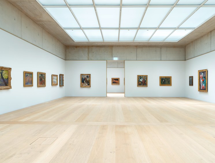 Modern gallery interior with white walls displaying framed paintings and natural lighting from glass ceiling