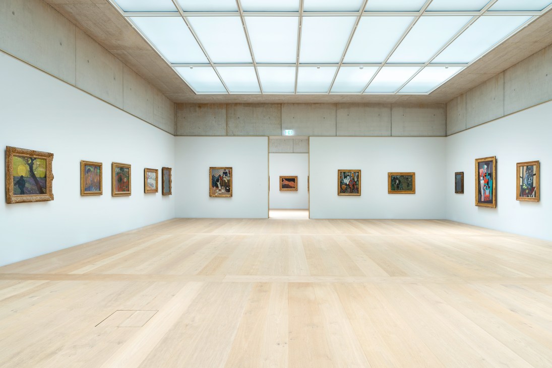 Modern gallery interior with white walls displaying framed paintings and natural lighting from glass ceiling