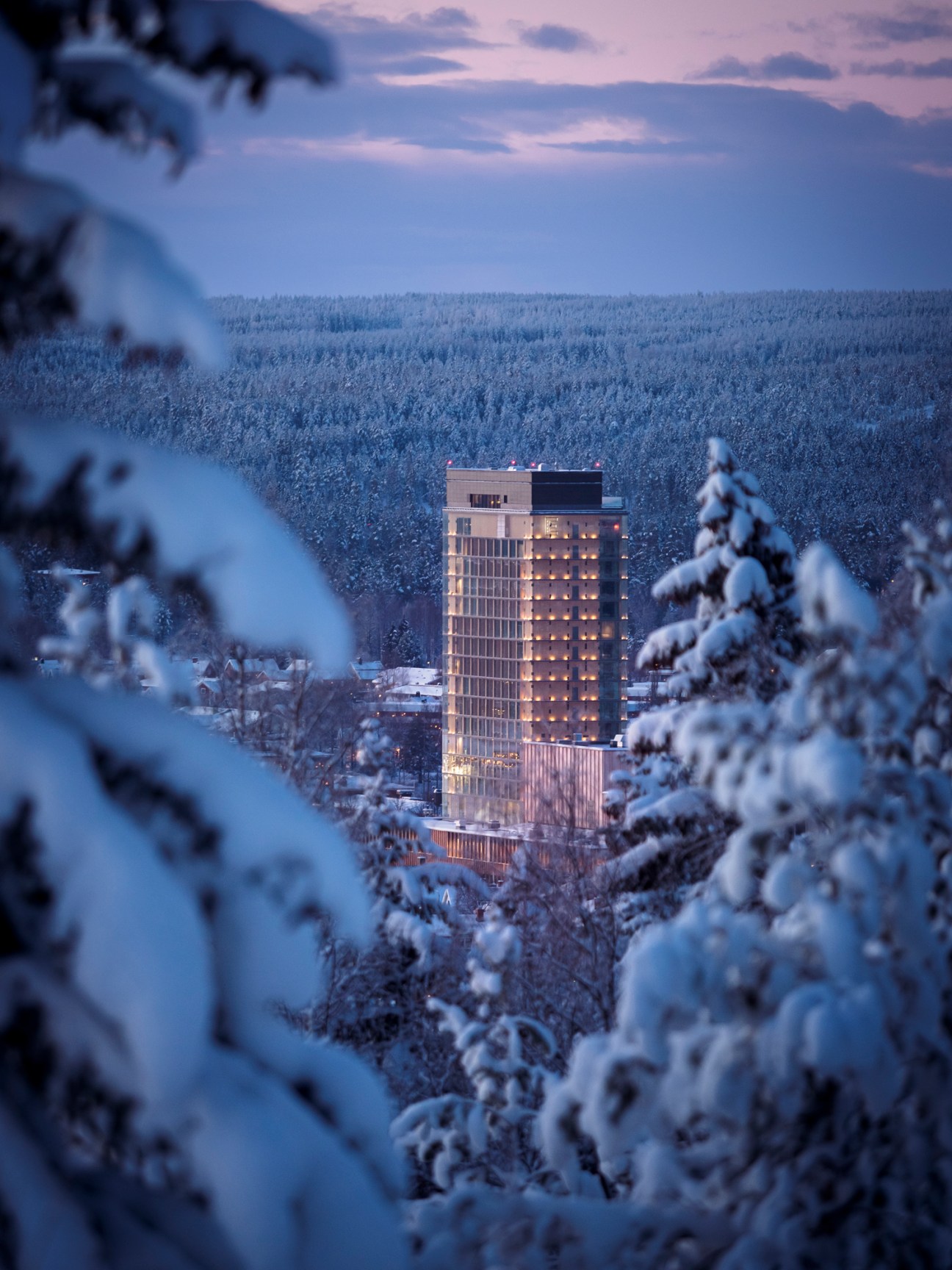 The Wood Hotel, a tall illuminated wooden tower building surrounded by snow-covered forest at dusk