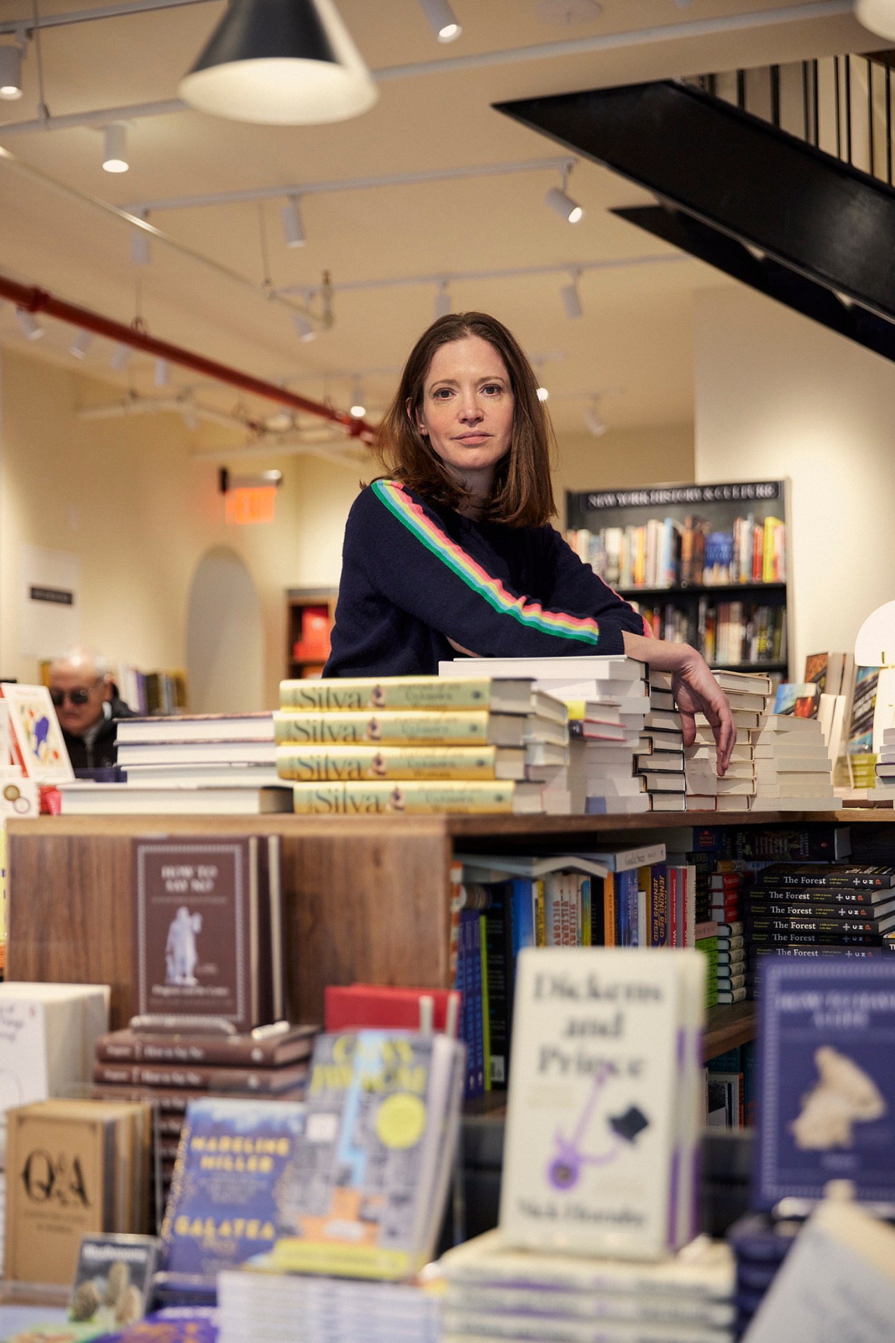 Woman with brown hair wearing dark sweater with rainbow stripe standing among stacks of books in bookstore