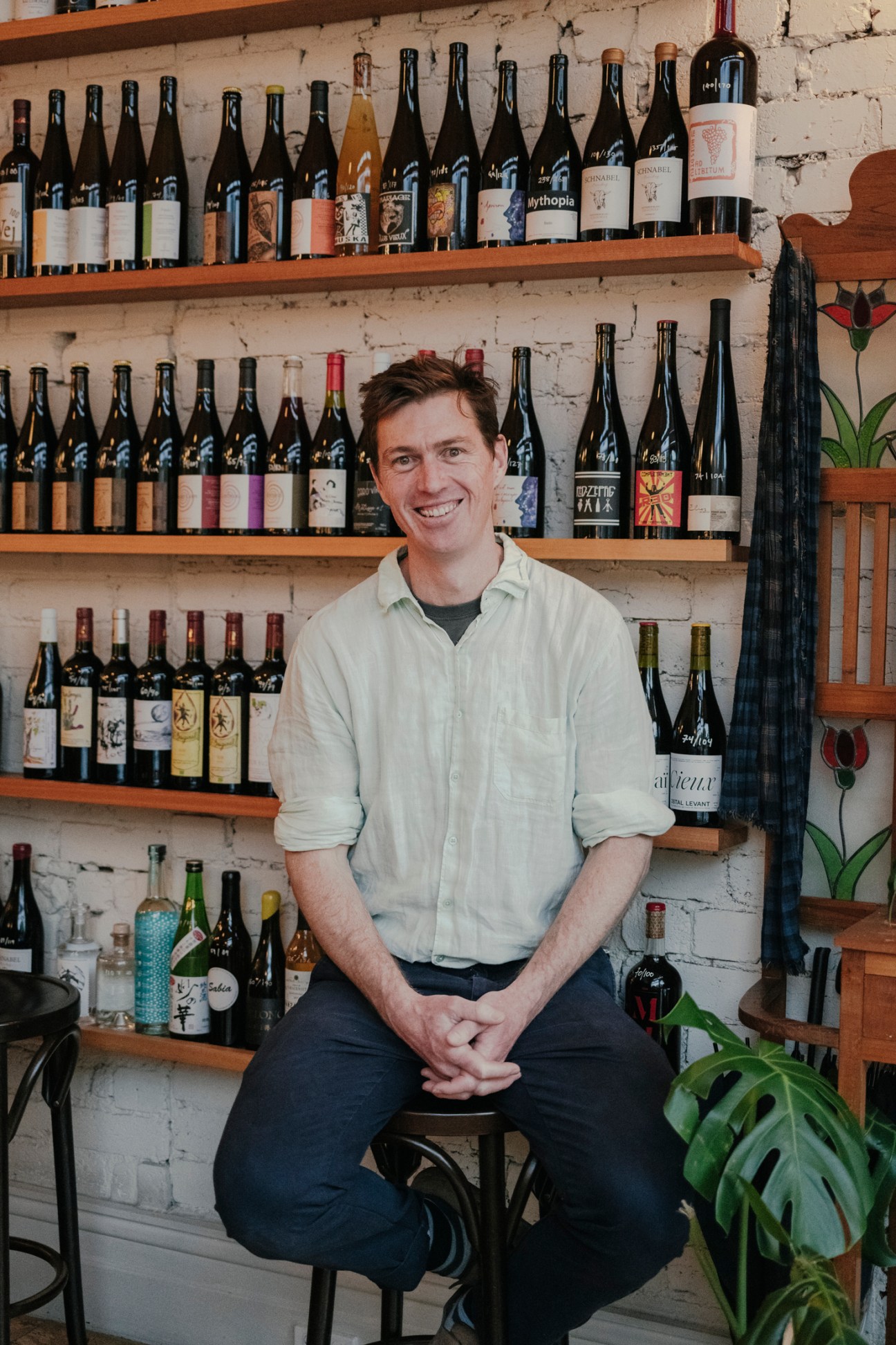 Man in light blue shirt sitting on stool surrounded by wine bottles on shelves
