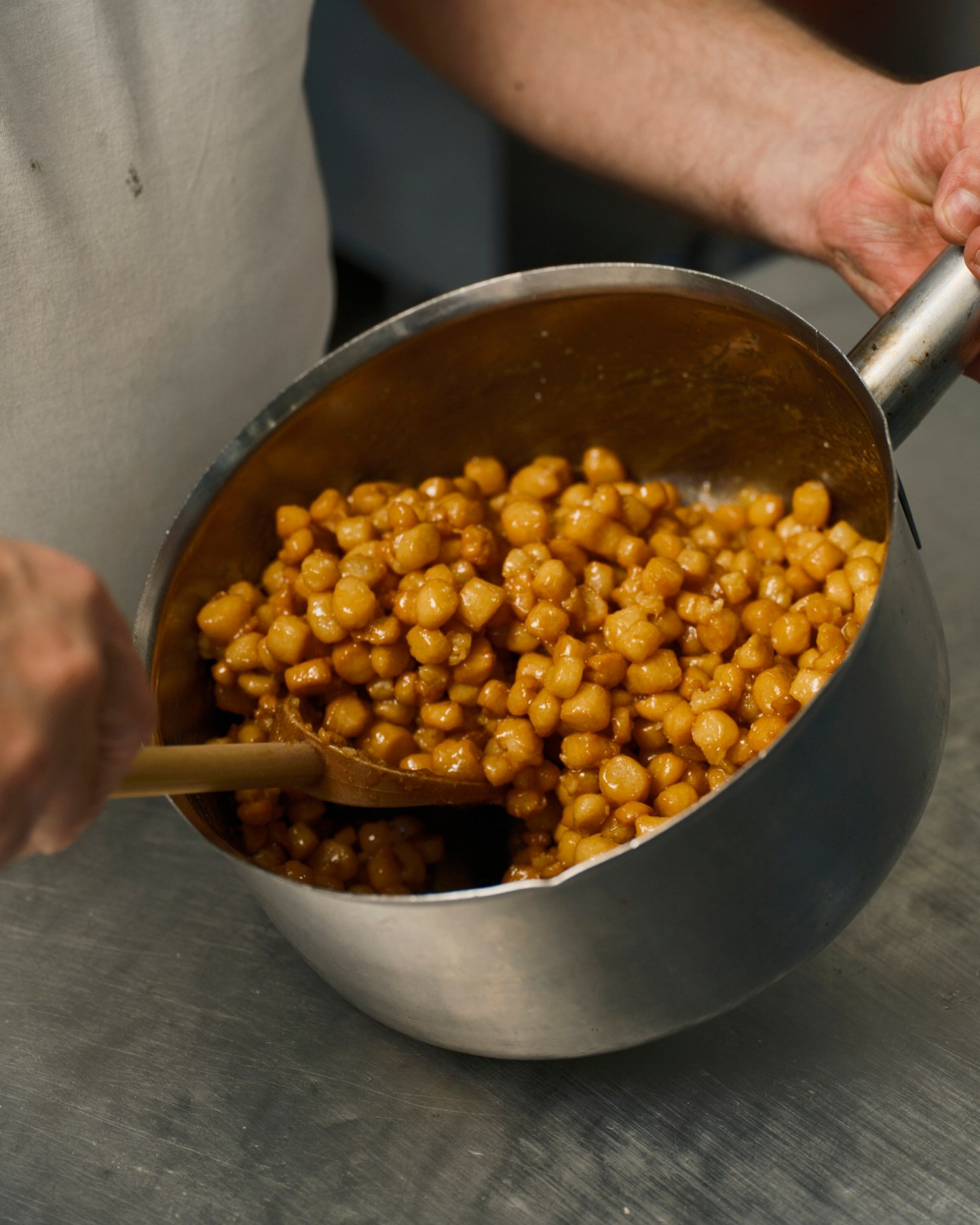 Hand holding a metal pot filled with golden honey-glazed struffoli balls.