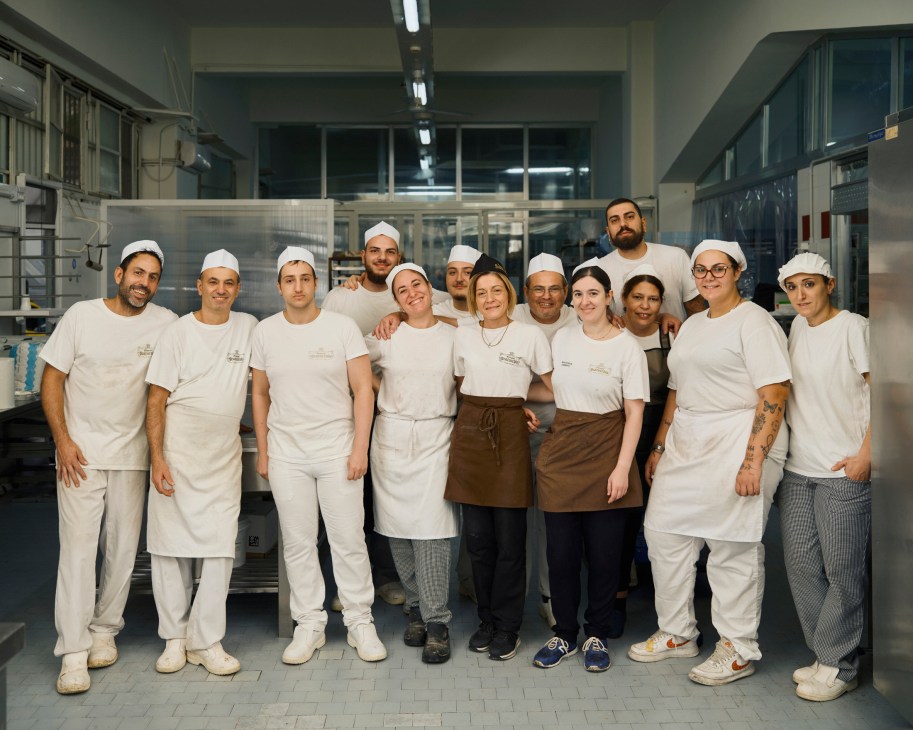 A group of pastry chefs and staff wearing white uniforms and hairnets pose together in a modern bakery kitchen at Scaturchio in Naples.