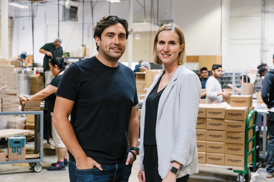 Jaime Schmidt and Michael Cammarata stand in a warehouse surrounded by cardboard boxes of Schmidt's Naturals products.