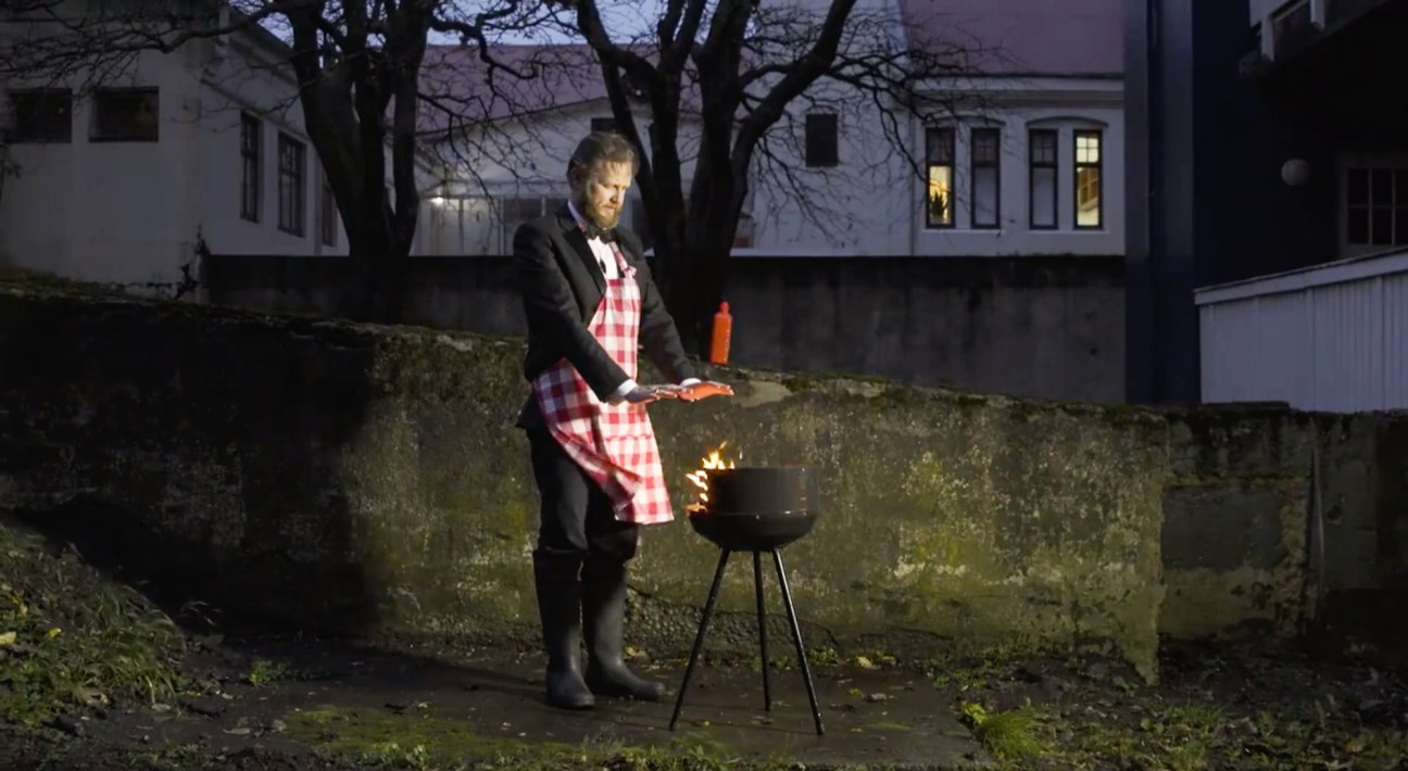Man in formal attire with checkered apron grilling outdoors at night near stone wall