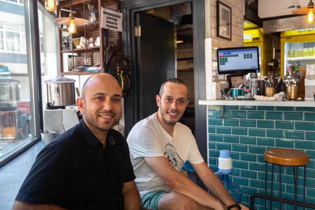 Two men sitting together in a modern restaurant with teal subway tile walls and industrial lighting