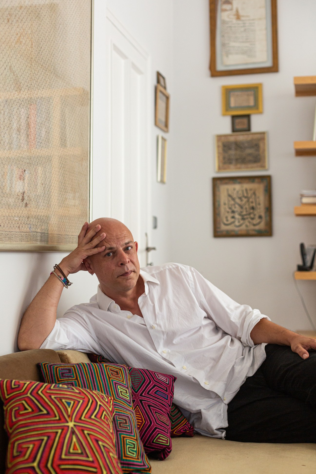 Portrait of gallerist Kerimcan Guleryuz sitting on a couch in a white shirt with framed artwork on the wall behind him