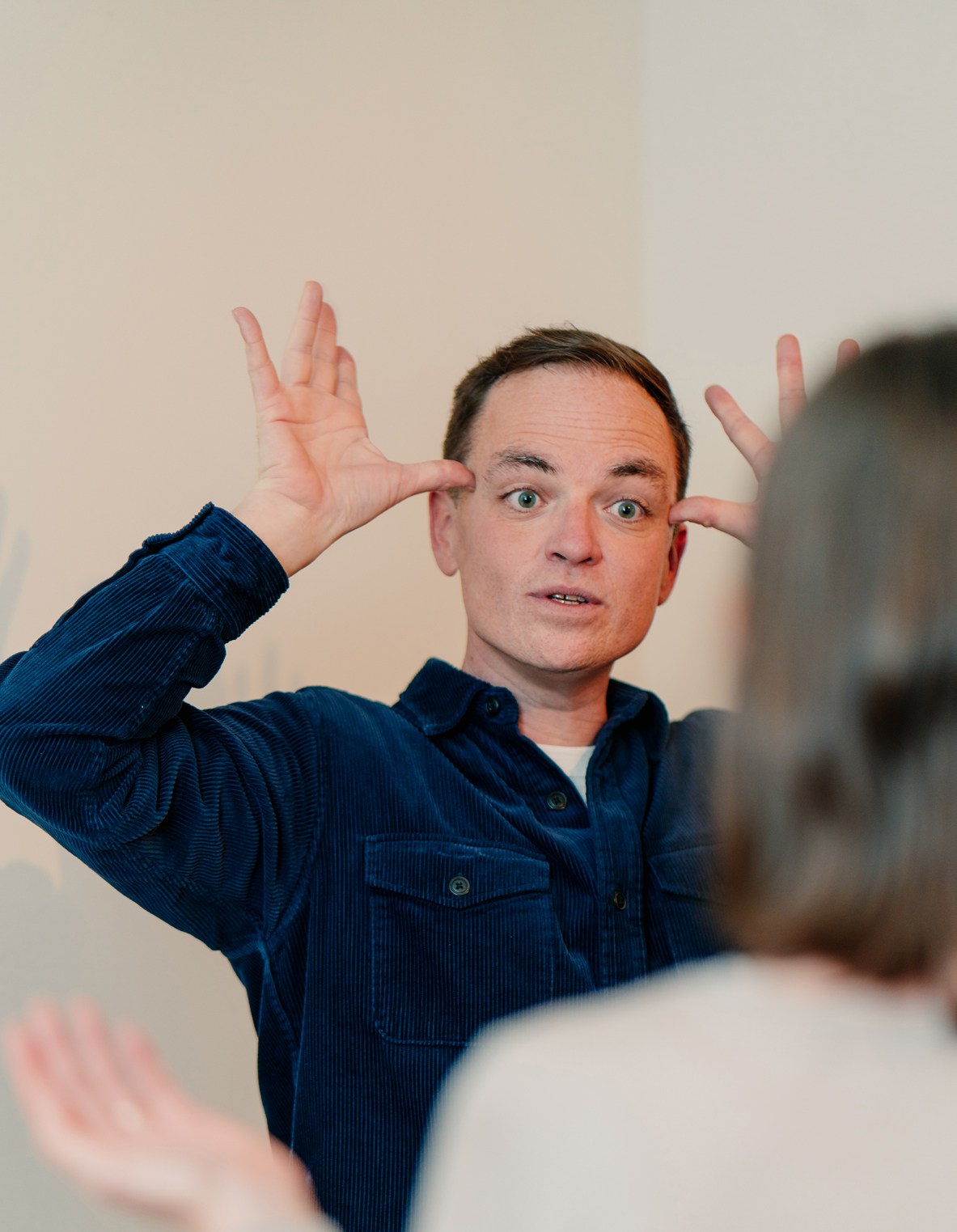 Man in blue corduroy shirt making exaggerated facial expressions with hands raised during improv class