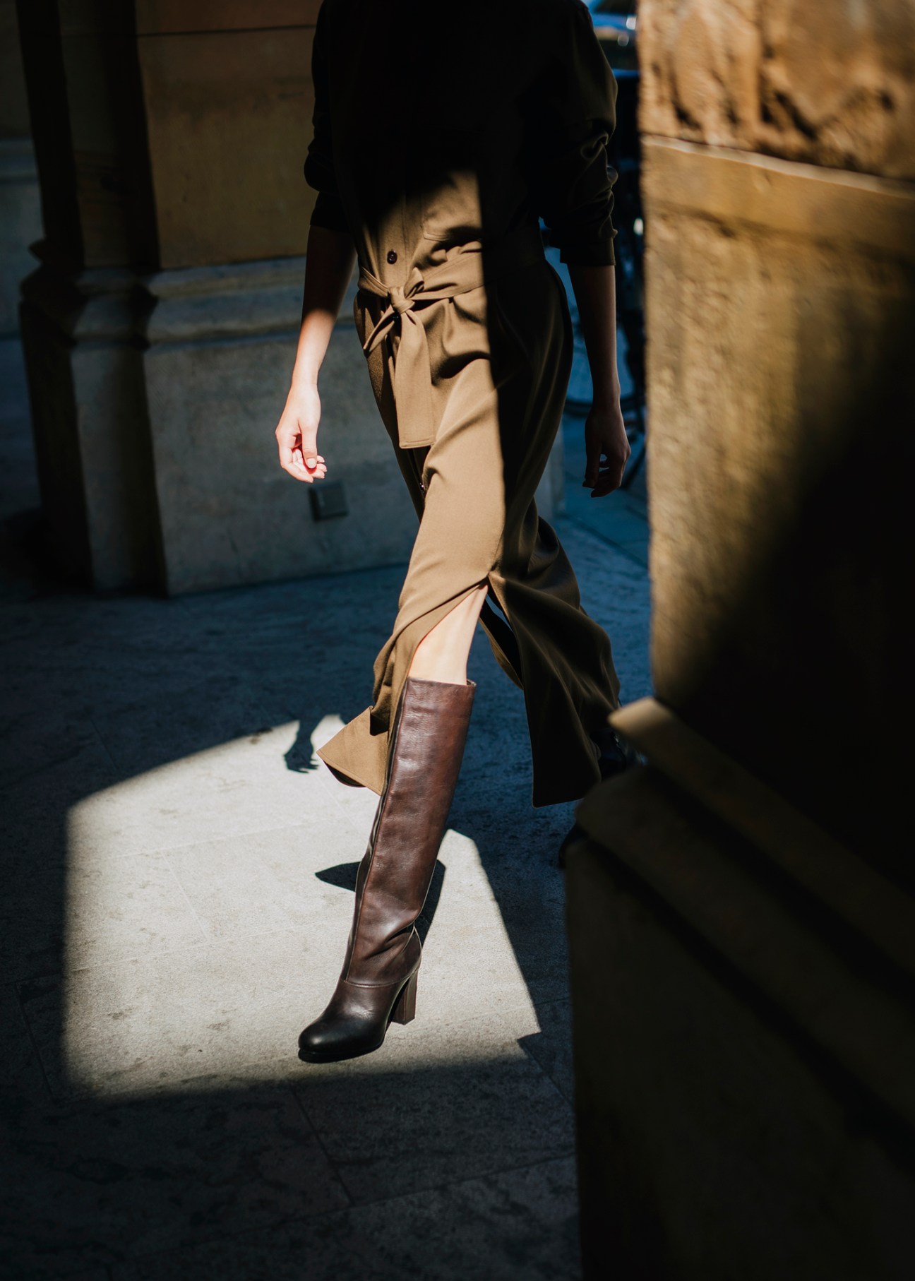 Person in beige trench coat and brown knee-high boots walking on cobblestone street in dramatic lighting