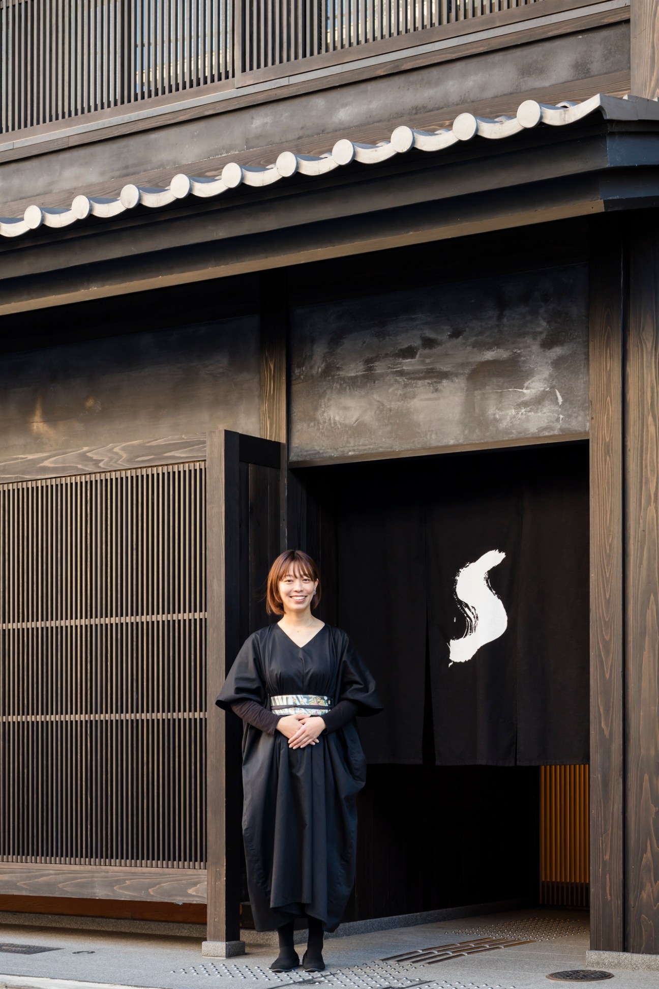 Woman in black kimono standing in front of traditional Japanese building entrance with noren curtain