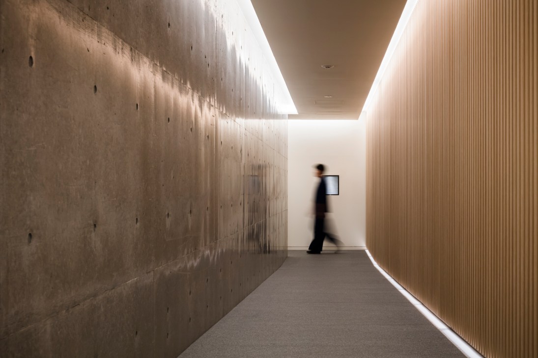 A blurred figure walks through a modern minimalist corridor with concrete walls and wooden paneling