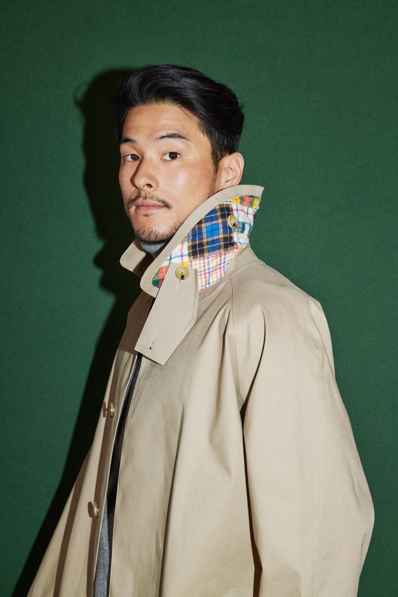 Man in beige Mackintosh coat with colorful plaid collar detail and Tod's rollneck jumper against green background