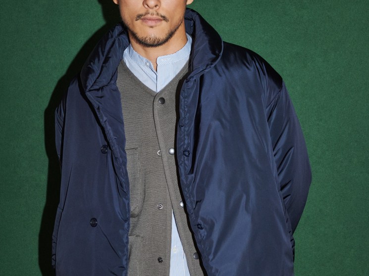 Man in layered autumn outfit featuring navy jacket over cardigan and shirt with glasses against green background