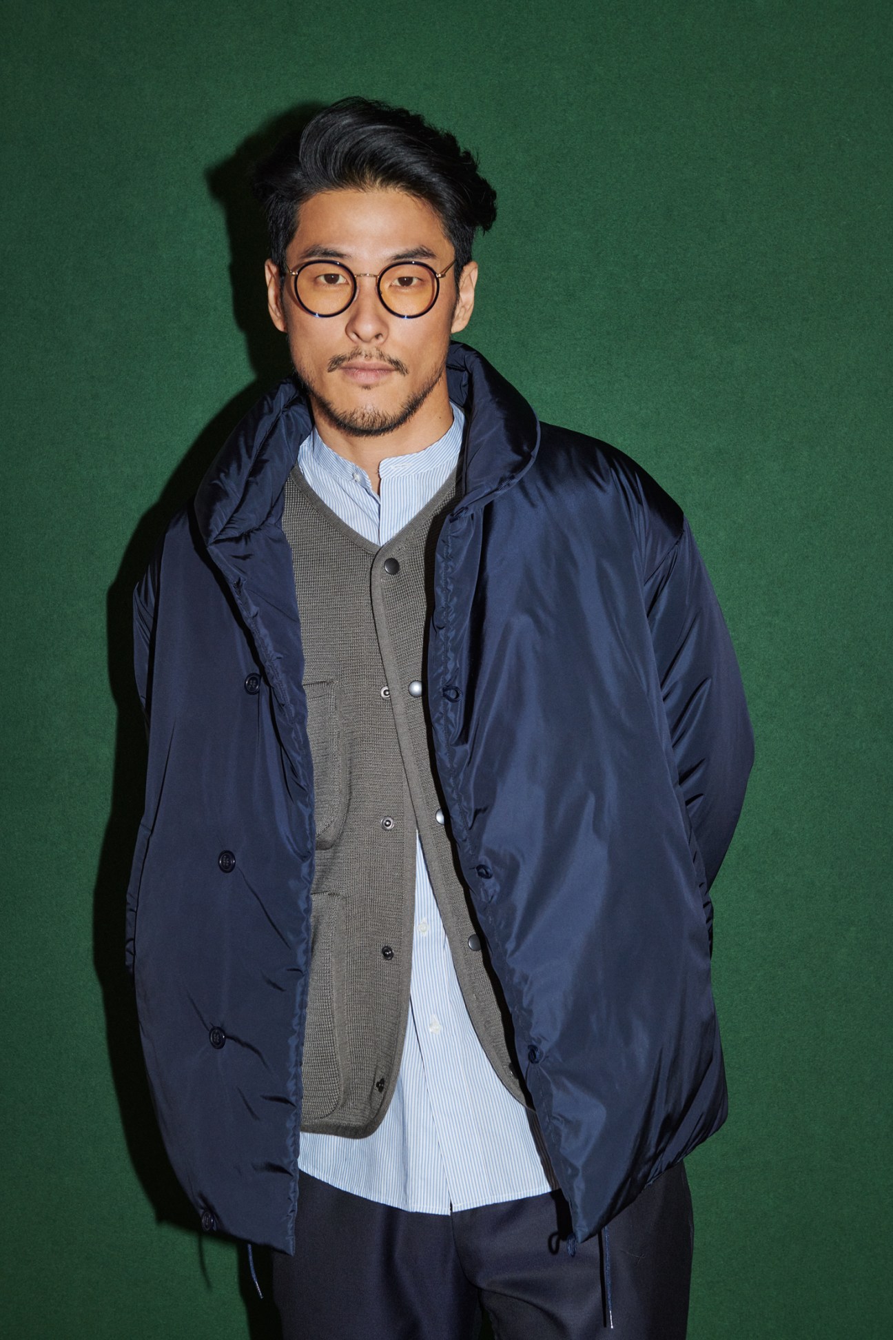Man in layered autumn outfit featuring navy jacket over cardigan and shirt with glasses against green background