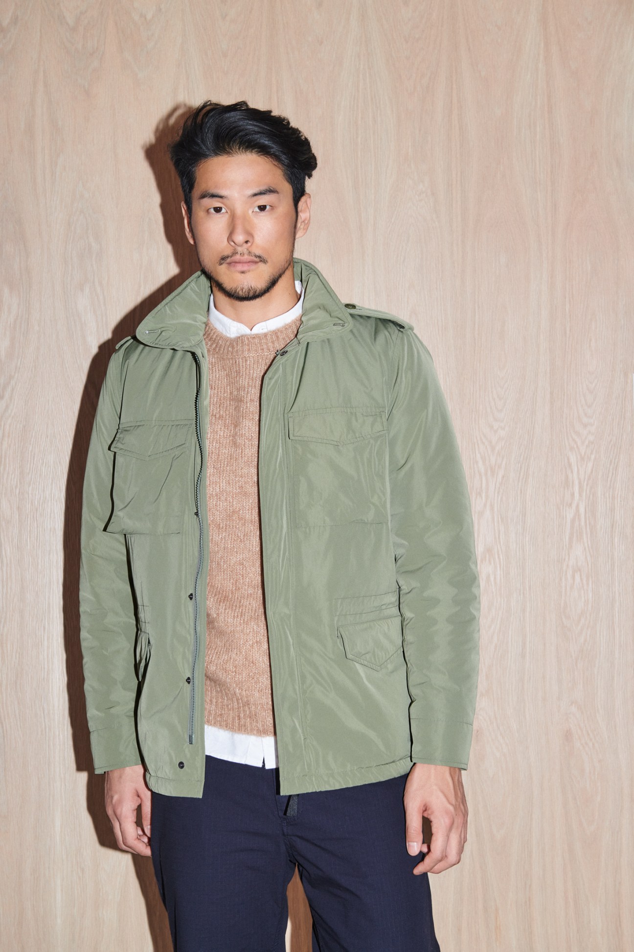 Man in olive green jacket over beige jumper and white shirt with dark trousers