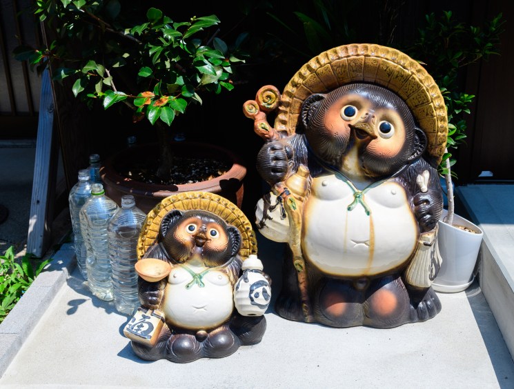 Two traditional Japanese tanuki (raccoon dog) ceramic figurines wearing straw hats displayed outside a building.