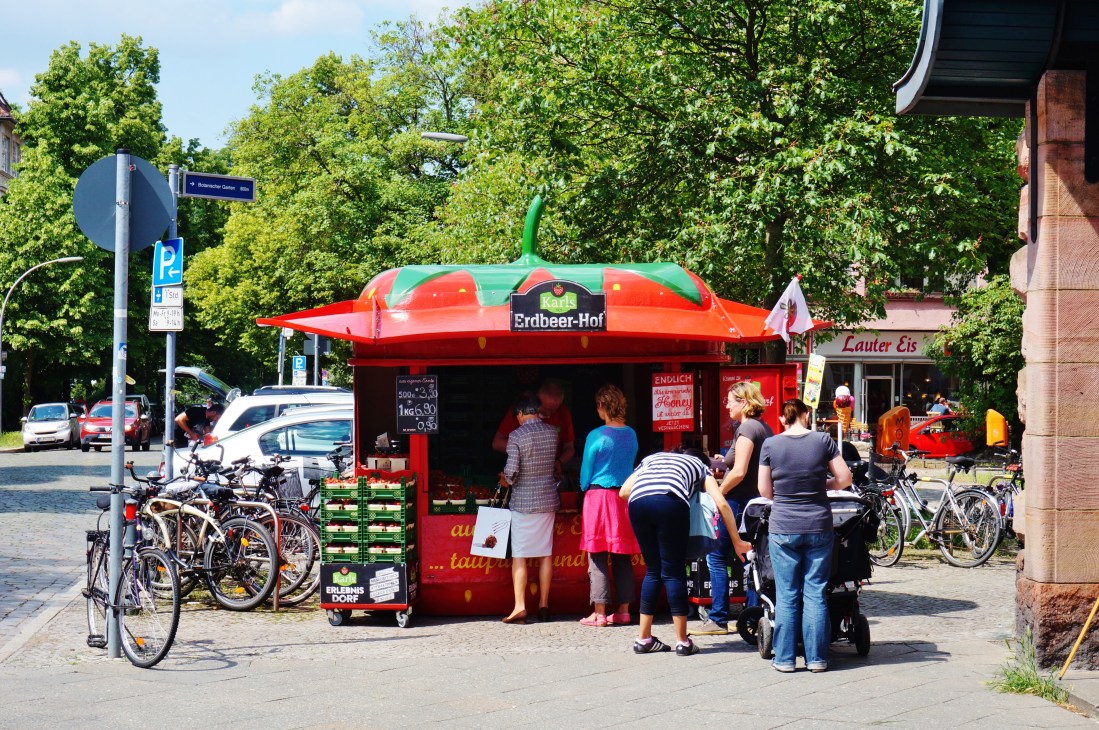 A red strawberry-shaped Karls Erdbeer-Hof booth with green stem on a Berlin street, surrounded by customers and bicycles.