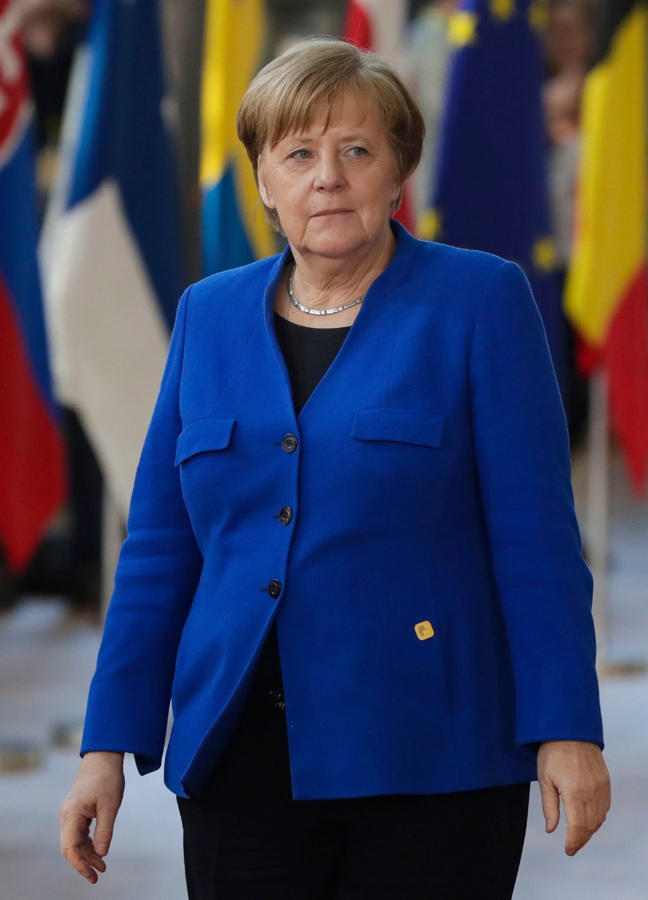 Angela Merkel wearing her characteristic blue blazer walking past European flags