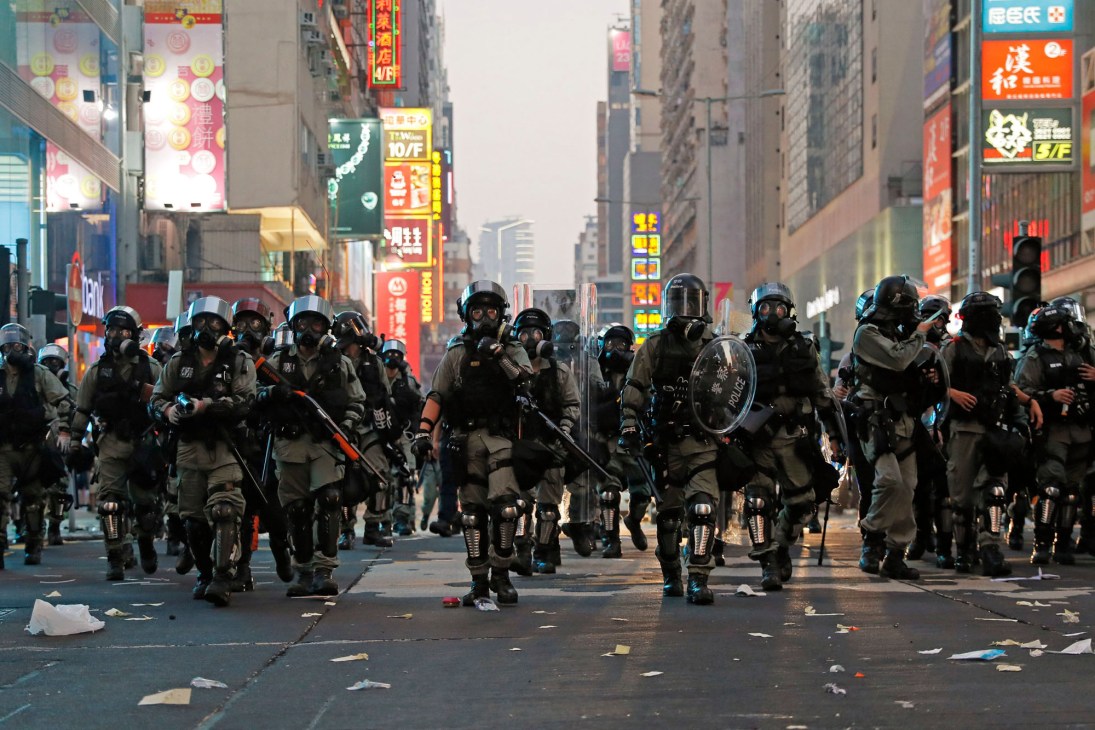 Riot police in tactical gear marching in formation down a Hong Kong street with colorful signage