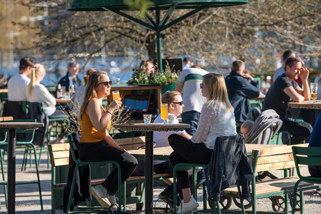 People seated at outdoor tables with green chairs and umbrellas in a Stockholm public space during spring.