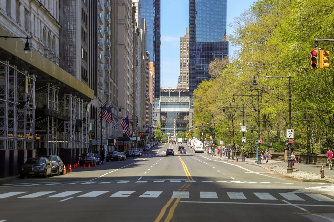 A quiet New York City street with green trees, traffic lights, and reduced pedestrian activity during pandemic lockdown.
