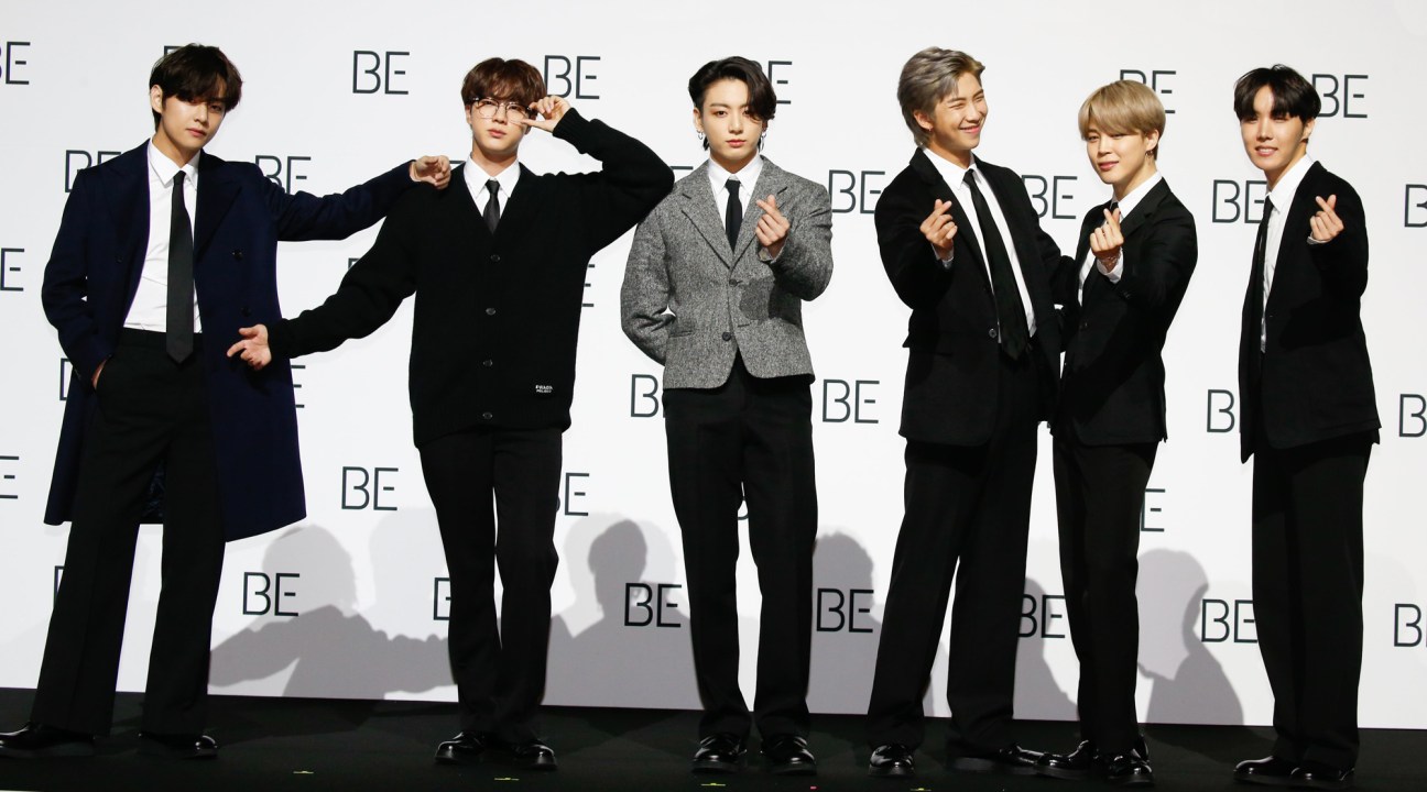 Six men in formal black and gray suits posing at a "BE" branded backdrop event