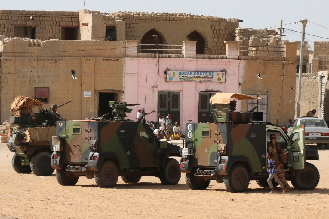 Two military armored vehicles with camouflage paint parked on dirt ground in front of mud brick buildings