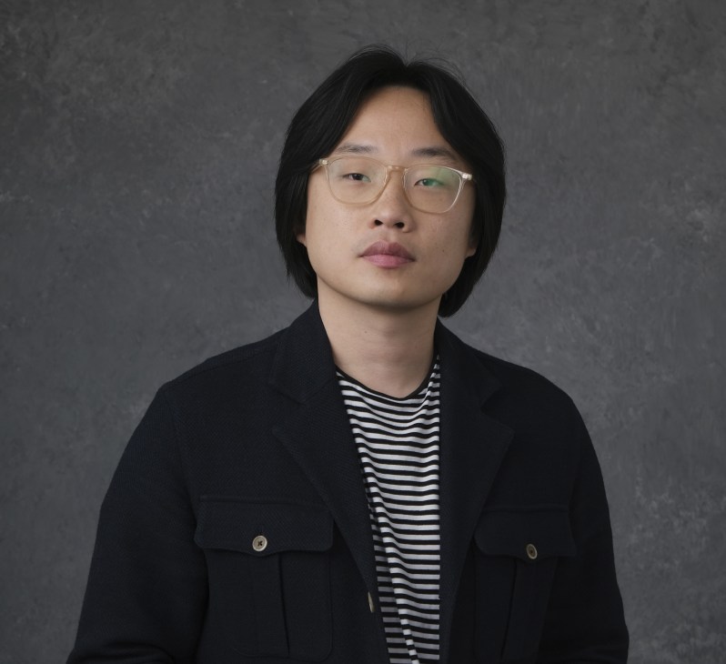 Jimmy O Yang wearing glasses and striped shirt under dark jacket against gray background