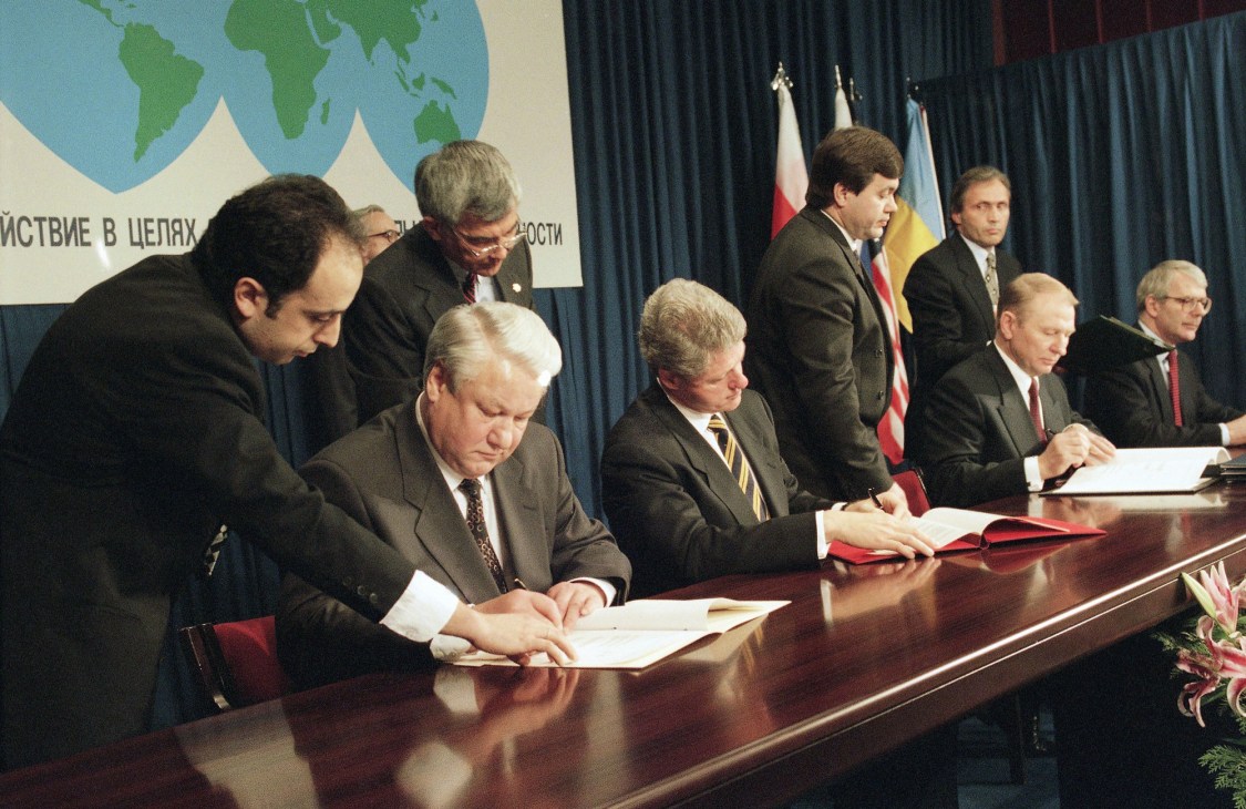 Officials signing documents at formal ceremony with flags and world map backdrop