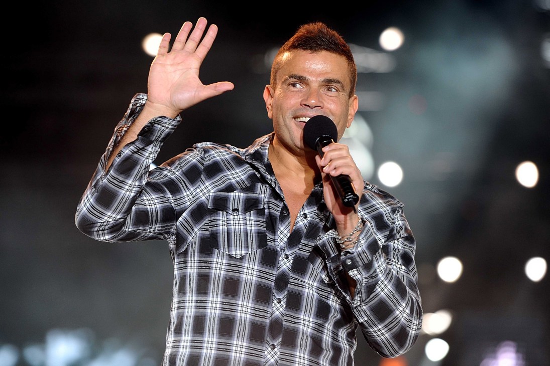 Amr Diab performing on stage with microphone, wearing plaid shirt and waving to audience