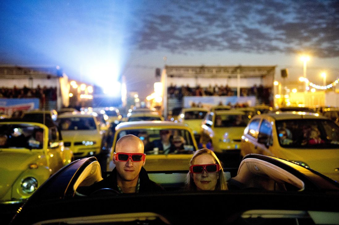 Two people wearing 3D glasses sit in a convertible car at a drive-in cinema with yellow cars and screens in background