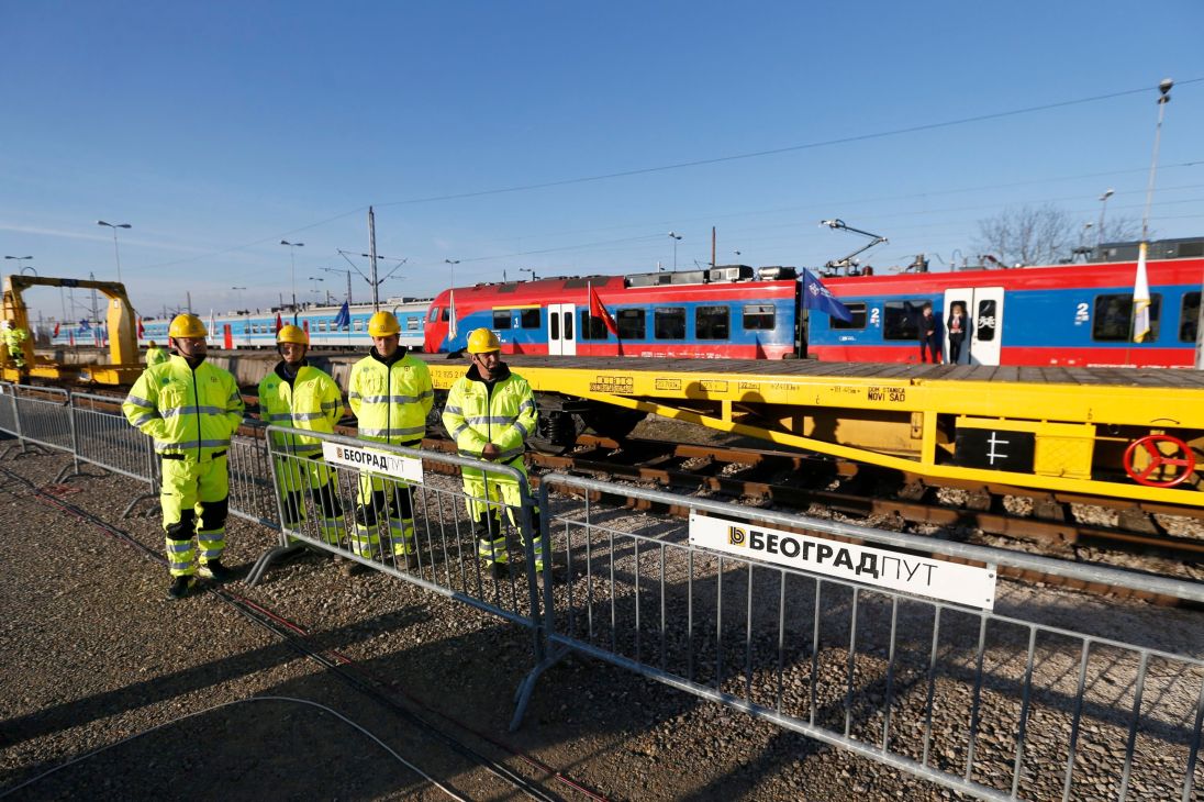 Construction workers in high-visibility gear stand beside yellow railway maintenance equipment and Belgrade-Budapest railway line tracks.