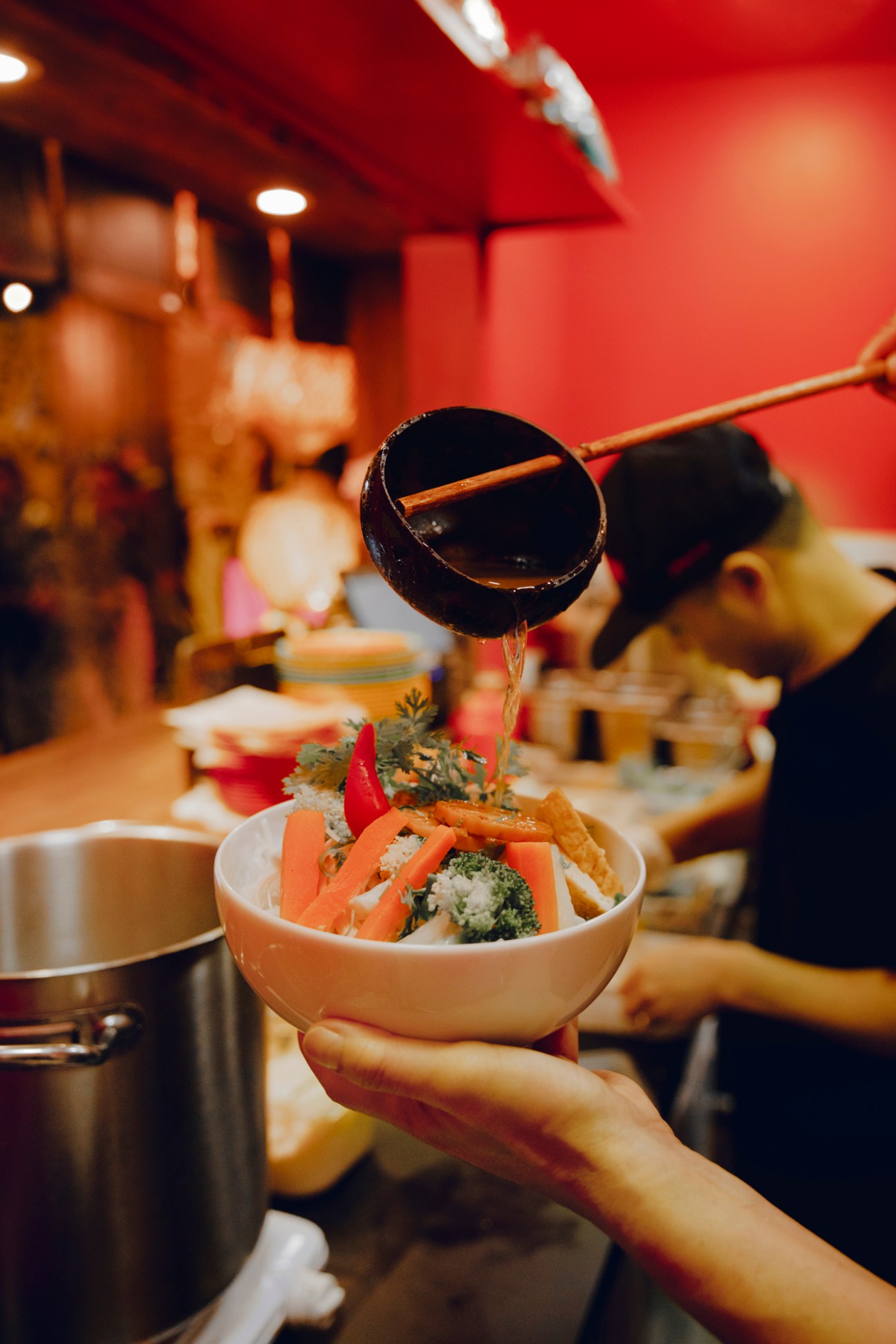 Hand holding a bowl of colorful Asian noodle dish with vegetables and dark sauce being poured from ladle