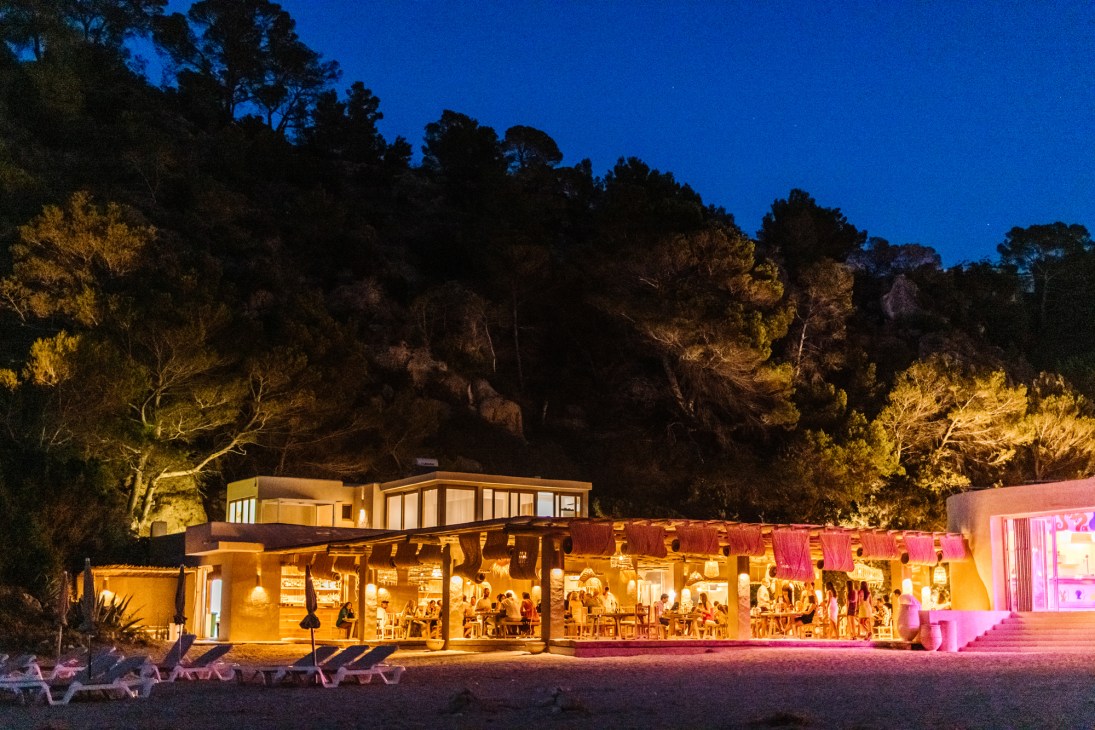El Silencio beach club on Ibiza illuminated at dusk with golden and pink lighting, surrounded by pine trees and sandy beach.