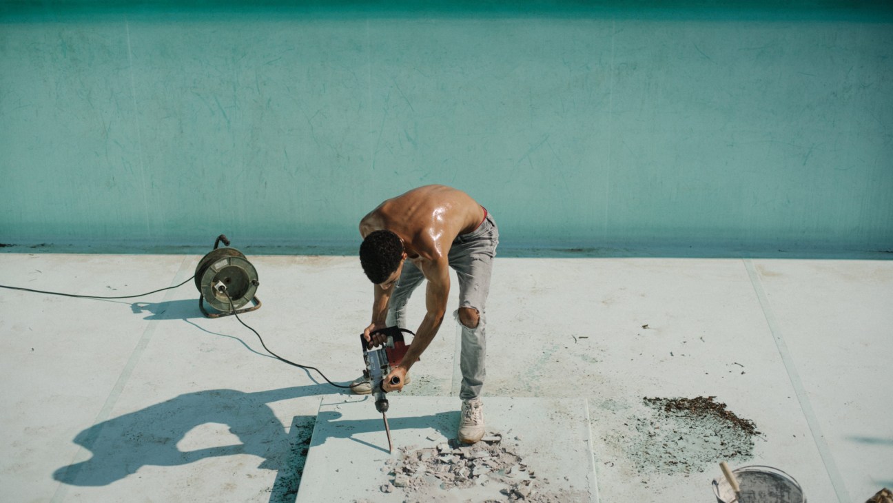 A shirtless man using a jackhammer on a white concrete surface near a turquoise wall