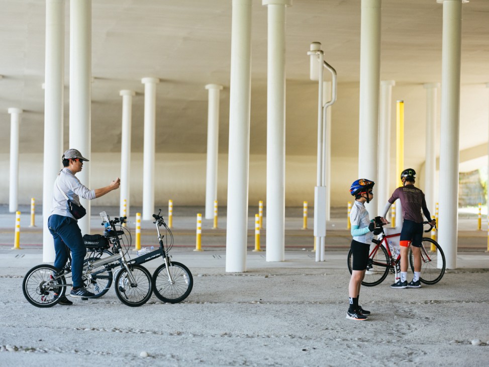 People with bicycles gathered under concrete pillars in an urban covered area