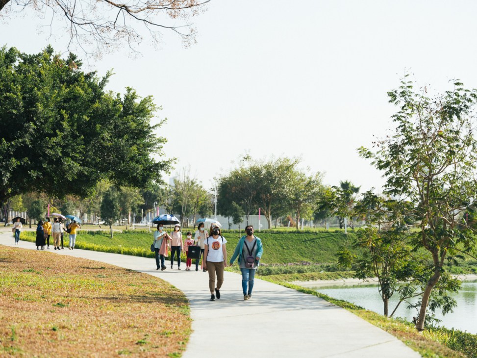 People walking along a paved path beside water with trees and green spaces